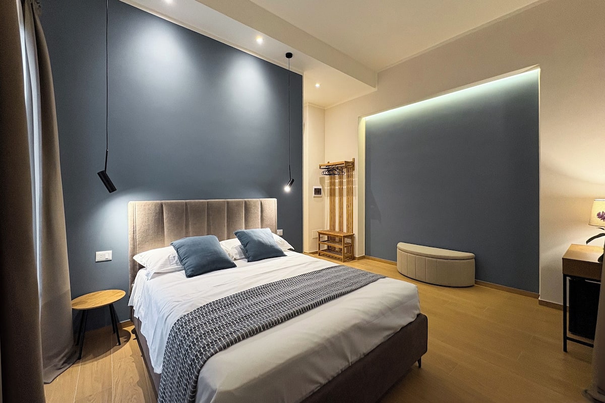 Moderni makuuhuone, jossa on suuri sänky, yöpöydät, pieni pyöreä pöytä ja seinään kiinnitetty valaisin.