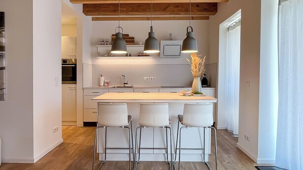 A modern kitchen with a central island, bar stools, pendant lights, and a wooden ceiling.