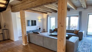 A spacious living area with a large sofa, a coffee table, and a TV mounted on the wall. There are wooden beams on the ceiling and a large window providing natural light.