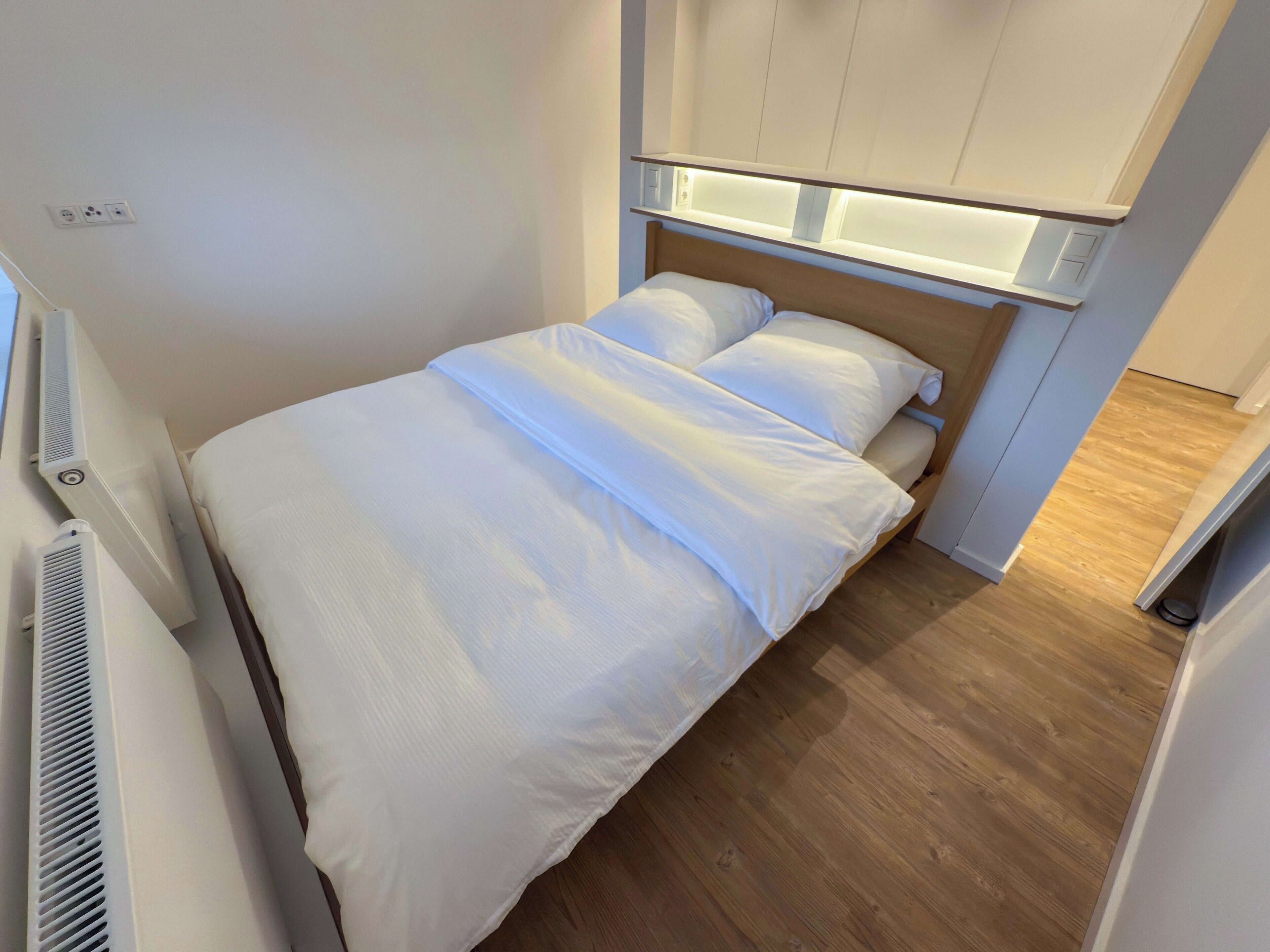 A compact bedroom with a bed, a built-in heater, and a wooden floor.