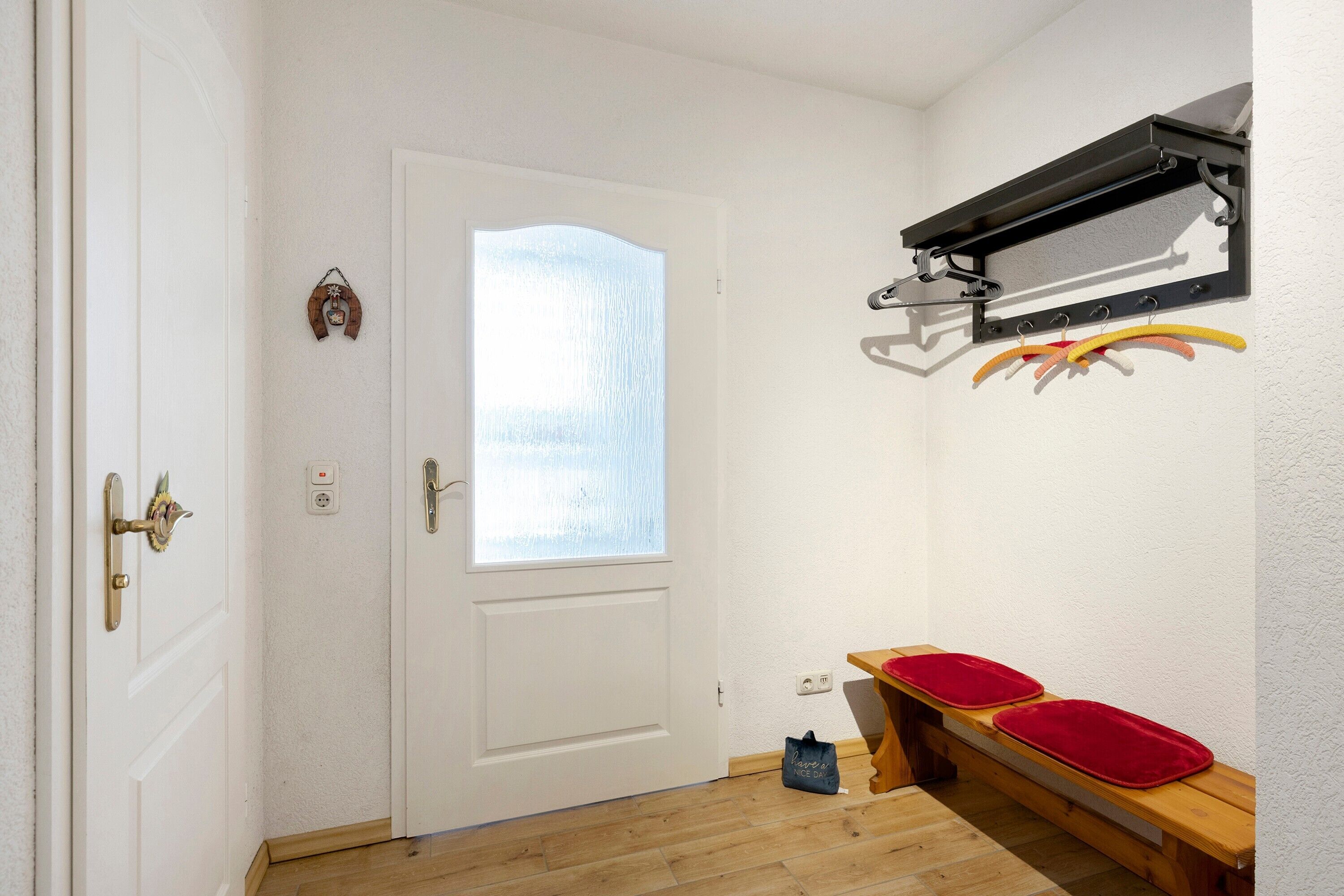 A room with a white door, a small window, a coat rack with hangers, and a wooden bench with red cushions.