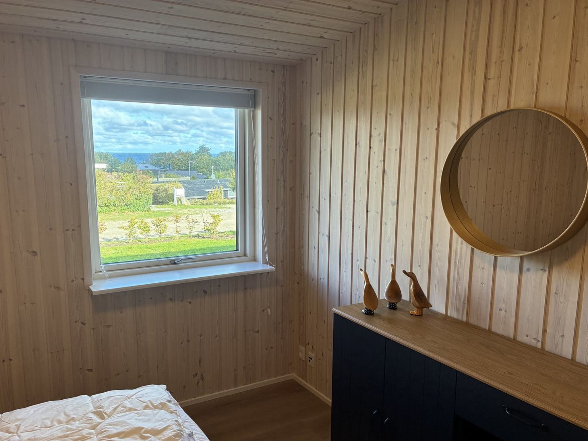 A bedroom with a window, wooden walls, a bed, a dresser, and a round mirror.