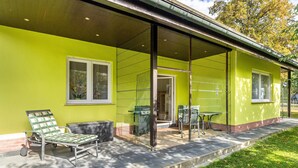 Een gebouw met een lichtgekleurde gevel, een overdekte veranda met een tafel en stoelen, en een gazon.