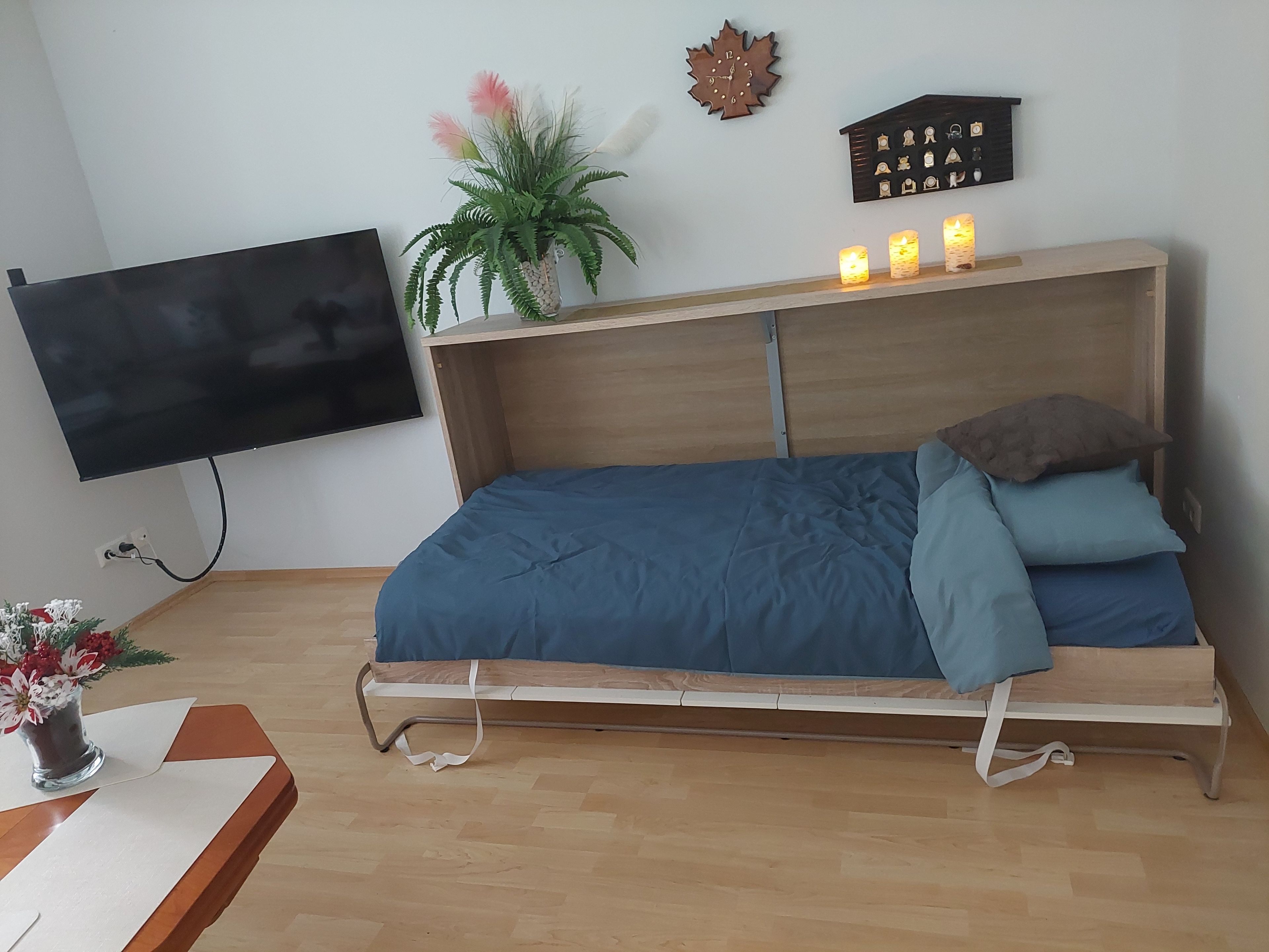 Une chambre à coucher avec un lit, un téléviseur fixé au mur, une plante et une étagère ornée d’objets décoratifs.