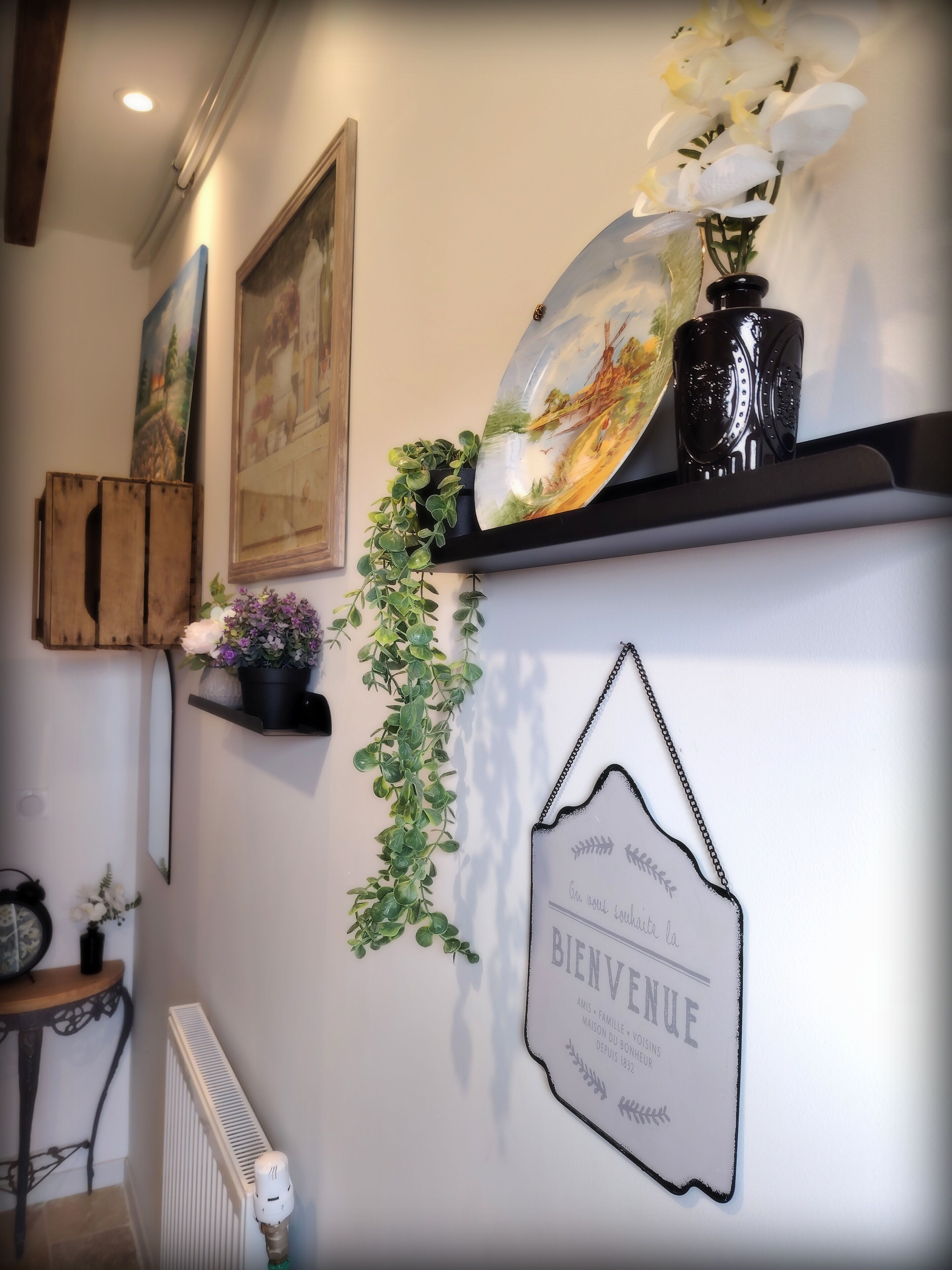 A wall with a welcome sign, a framed painting, and a shelf with a vase and flowers.