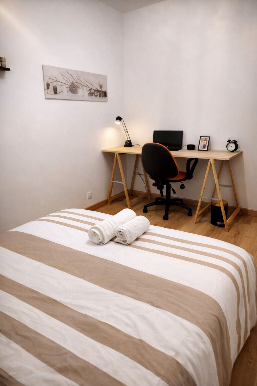 Una cama bien hecha con ropa de cama a rayas, un escritorio con una computadora, una silla y una mesita de noche con una lámpara y un reloj.