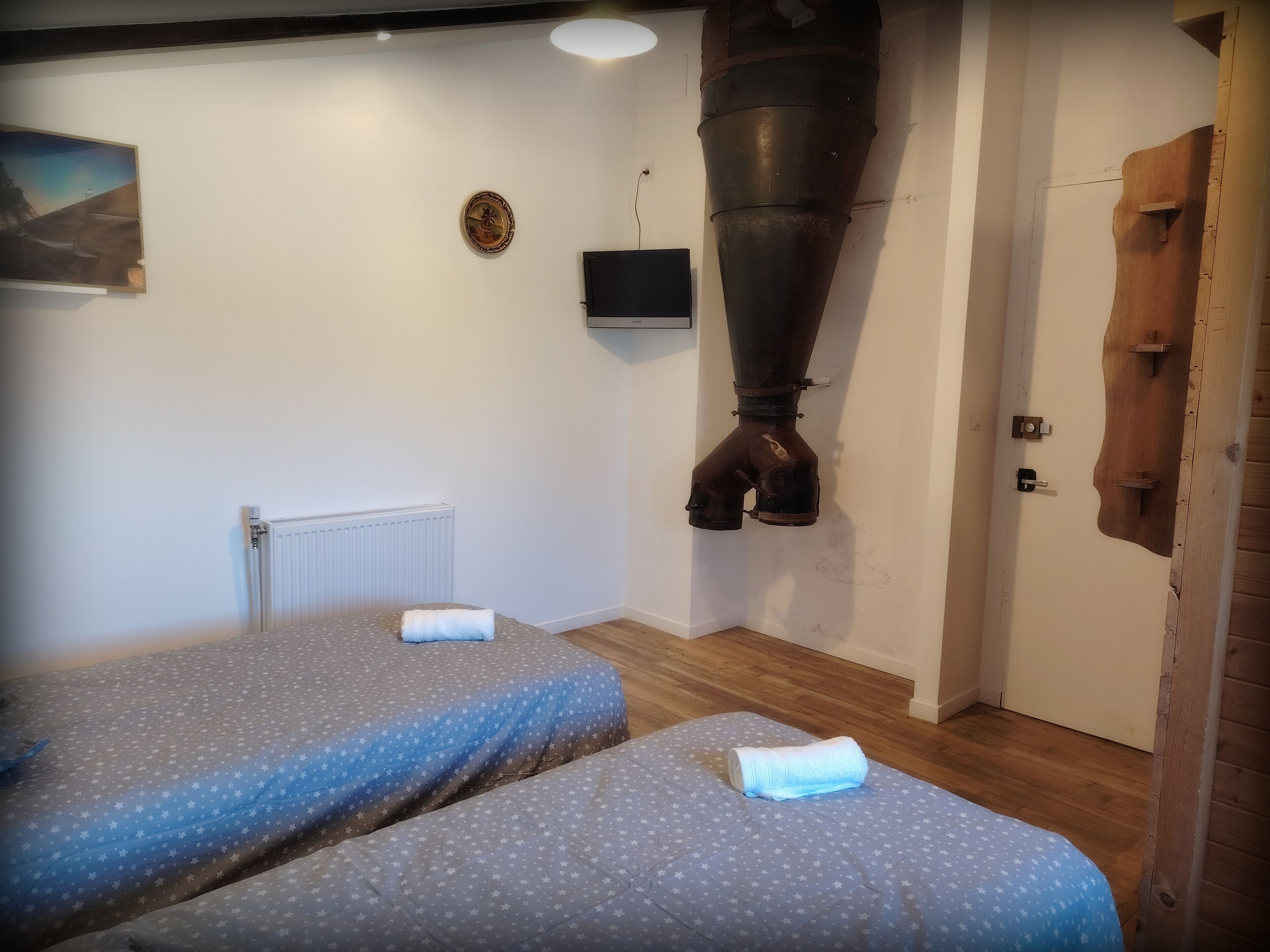 A room with two beds, a radiator, a large industrial fan, and a wooden door.