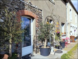 Um edifício rústico com fachada de pedra, porta de madeira e um pequeno jardim com vasos de plantas e móveis de exterior.
