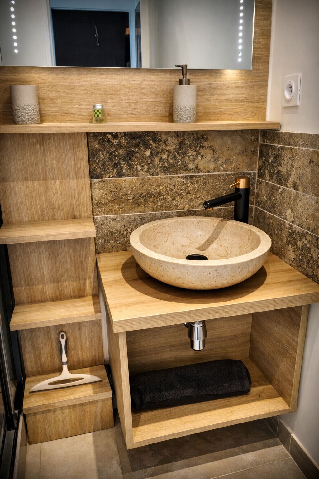 Un baño moderno con un lavabo redondo color beige, estantes de madera y una toalla negra.
