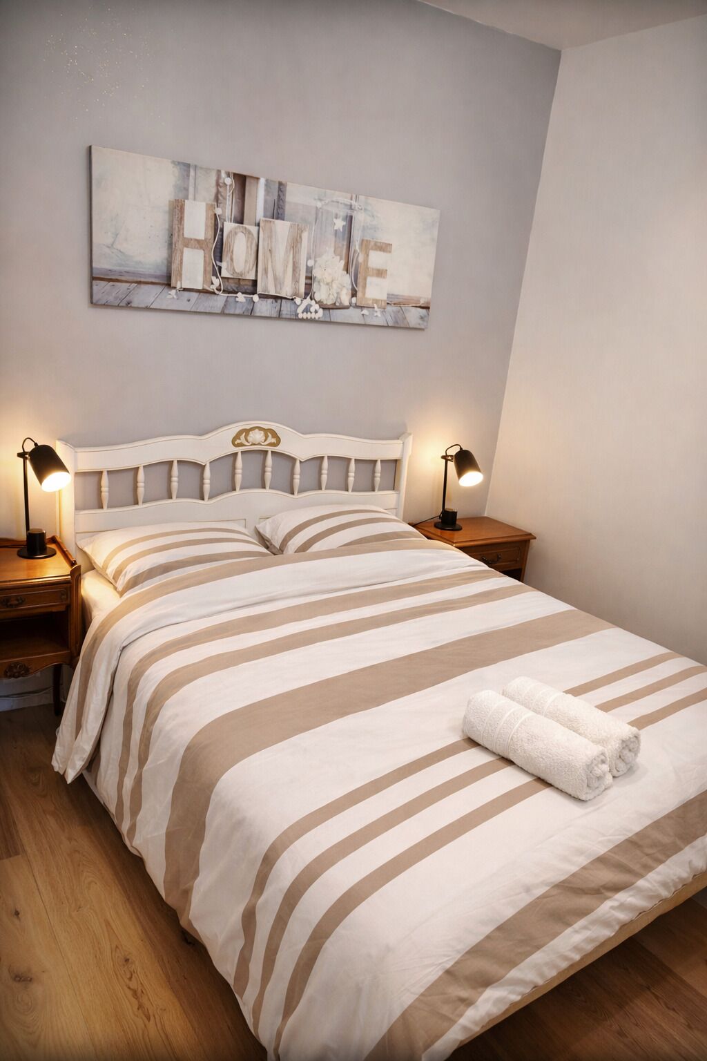 A neatly made bed with striped bedding, two bedside lamps, a wooden headboard, and a painting above the bed.