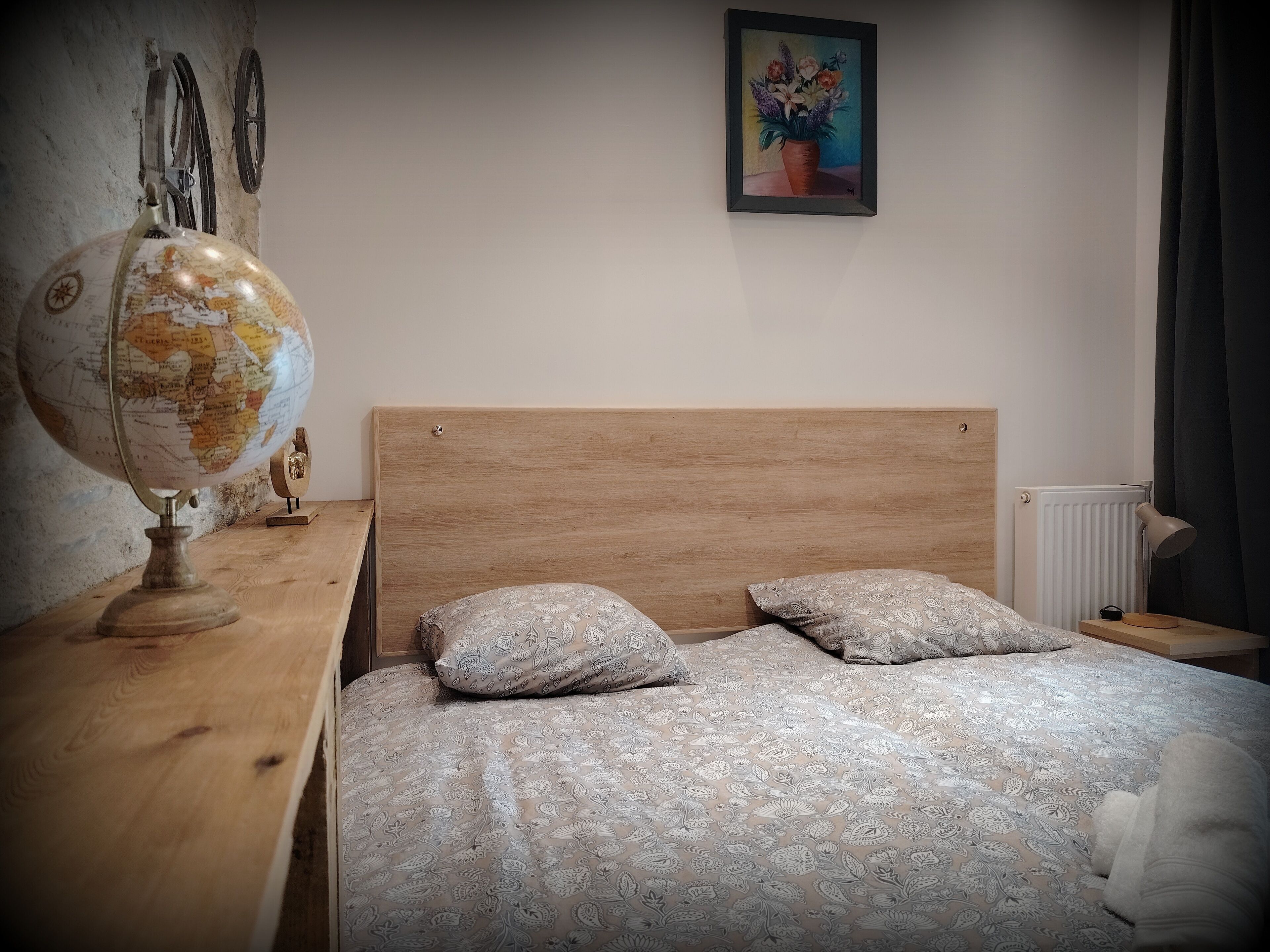 Un dormitorio con cabecera de madera, un globo terráqueo, almohadas estampadas, un cuadro y un radiador.
