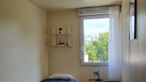 A small, well-lit room with a bed, a desk, and a chair. There is a window with curtains, a shelf with decorative items, and a ceiling light. - Résidence hôtelière ERASMUS (Malakoff)