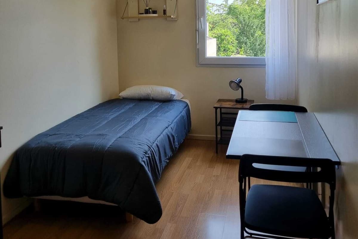 A small, well-lit room with a bed, a desk, and a chair. There is a window with curtains, a shelf with decorative items, and a ceiling light.