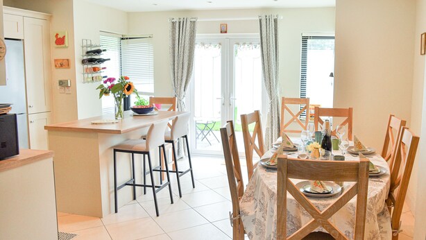A dining area with a table set for four, chairs, a kitchen area with a counter, and a view through a glass door. - Skellig Fuchsia Lodge Ballinskelligs (Ballinskelligs)