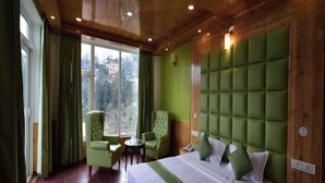 A hotel room with a large bed, green upholstered headboard, two armchairs, a small round table, and a view of the outdoors through large windows. - the town ville  (Shimla)