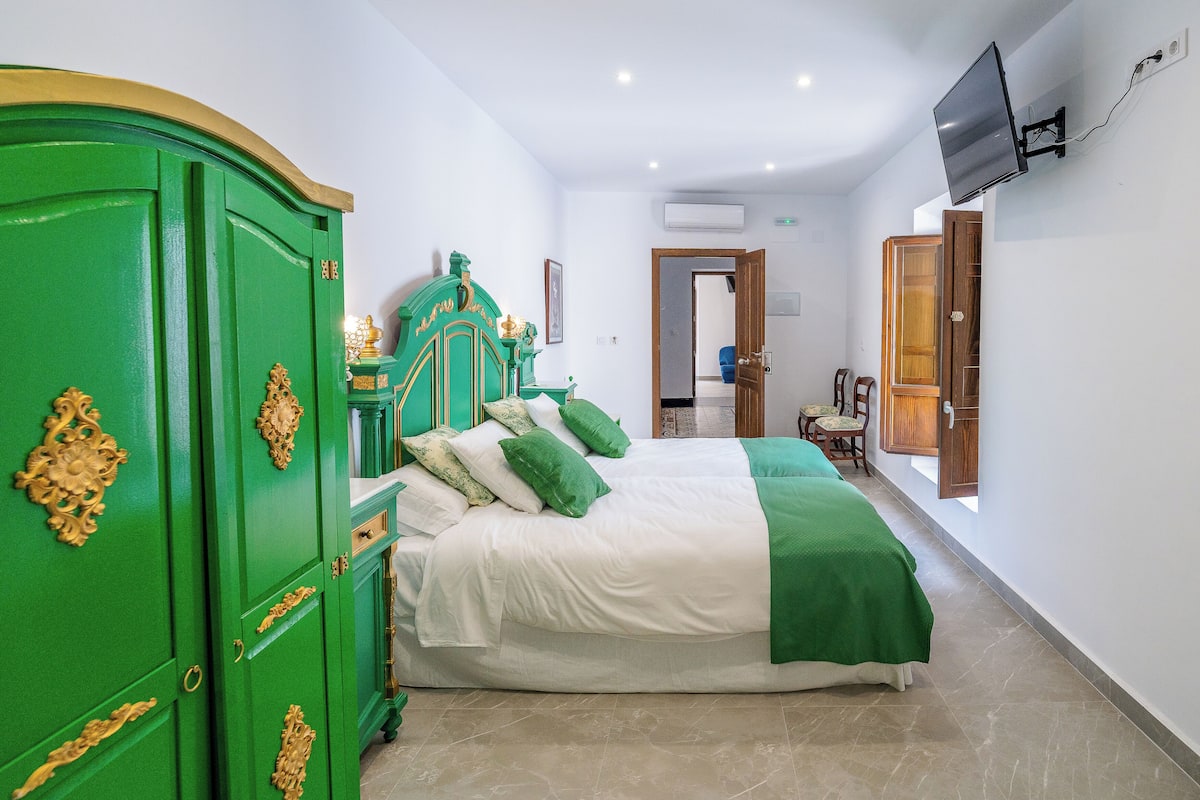 A hotel room with a green wardrobe, a bed with white and green bedding, a television mounted on the wall, and a wooden door.