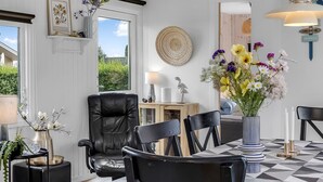 Un salon moderne avec un canapé en cuir noir, une table à manger avec des chaises noires et blanches, un vase rempli de fleurs et une grande fenêtre donnant sur un paysage verdoyant.