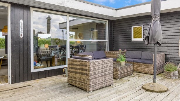 Une terrasse extérieure aménagée avec des meubles en osier, un parasol gris et une vue sur l’intérieur de la maison grâce à de grandes portes vitrées.
