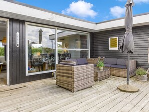 Een terras buiten met rieten meubilair, een grijze parasol en uitzicht op het huis door grote glazen deuren.