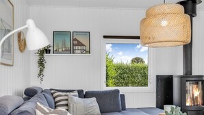 Un salon moderne avec un canapé gris, une table basse blanche, une lampe suspendue en osier, des photos encadrées du Danemark et un poêle à bois.