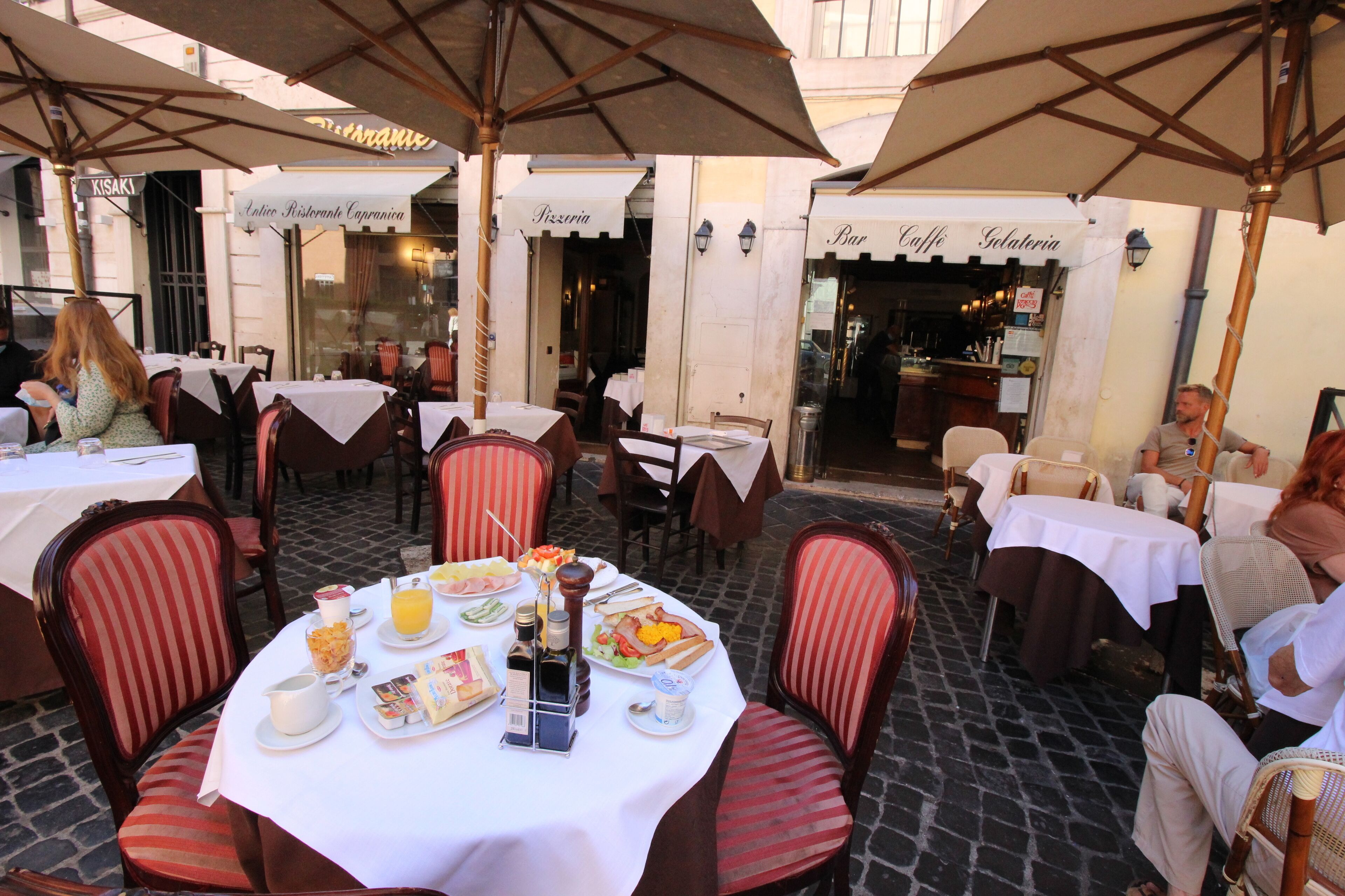 Café al aire libre con mesas, sillas, sombrillas y carteles de distintos establecimientos de comida y bebida.