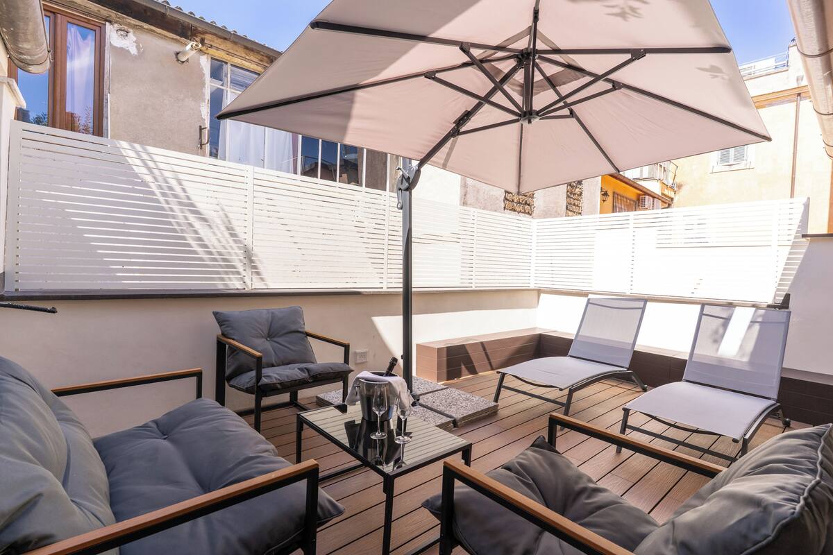A rooftop patio with a large umbrella, seating area, and a table with a bottle and glasses.