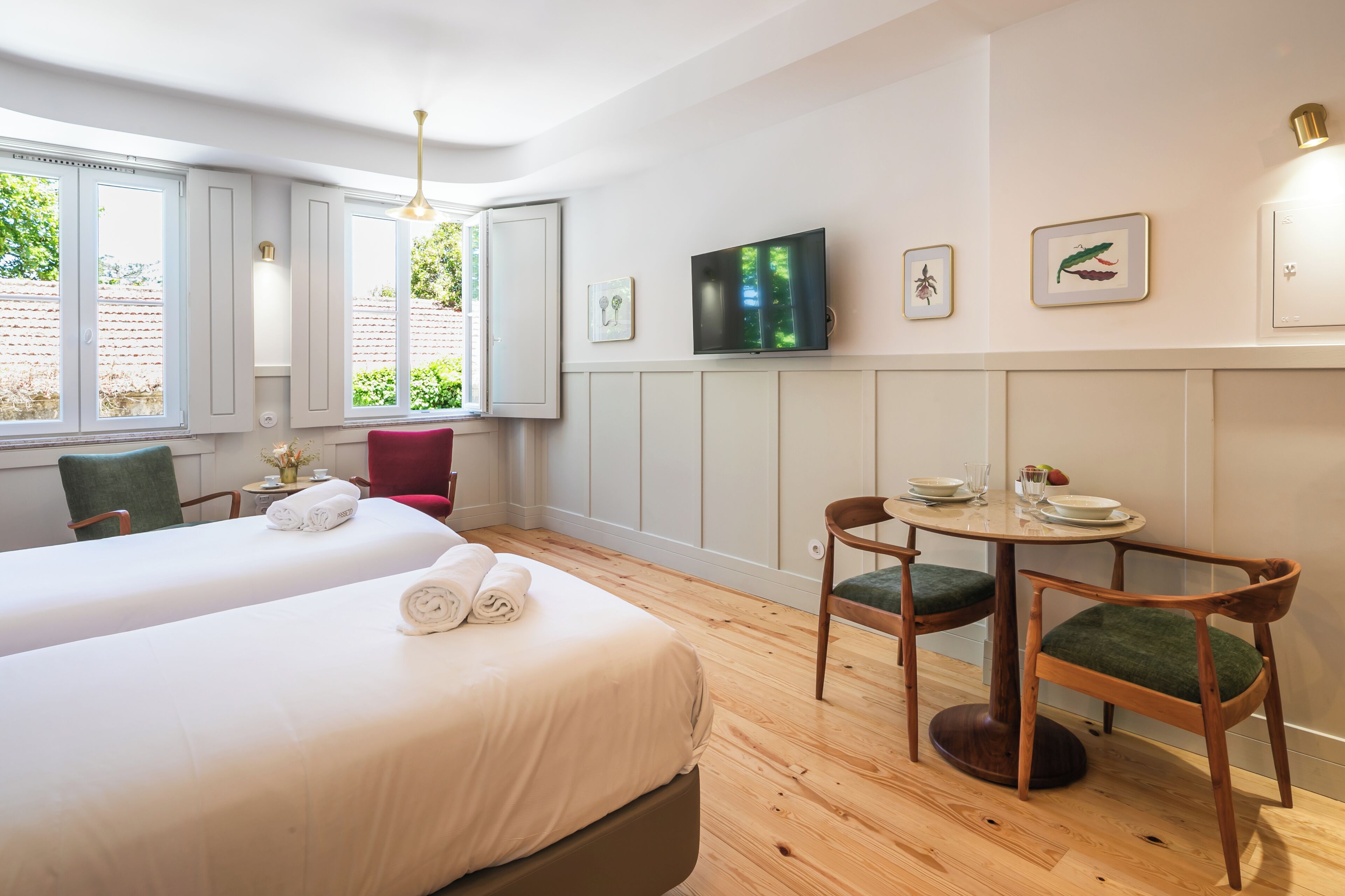 Habitación de hotel con dos camas, una mesa de comedor con sillas, un televisor montado en la pared y cuadros enmarcados.