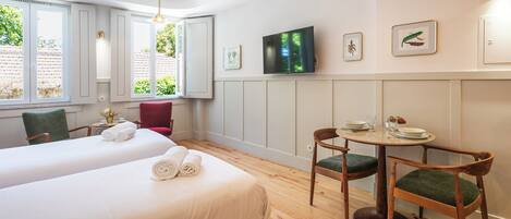 Habitación de hotel con dos camas, una mesa de comedor con sillas, un televisor montado en la pared y cuadros enmarcados.