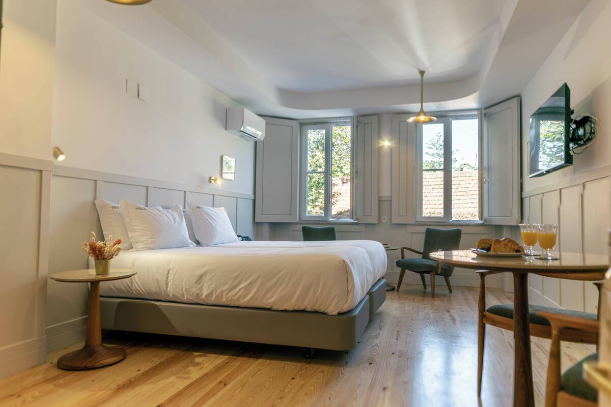 A bedroom with a bed, a small round table with a vase, a chair, and a breakfast table with croissants and orange juice.