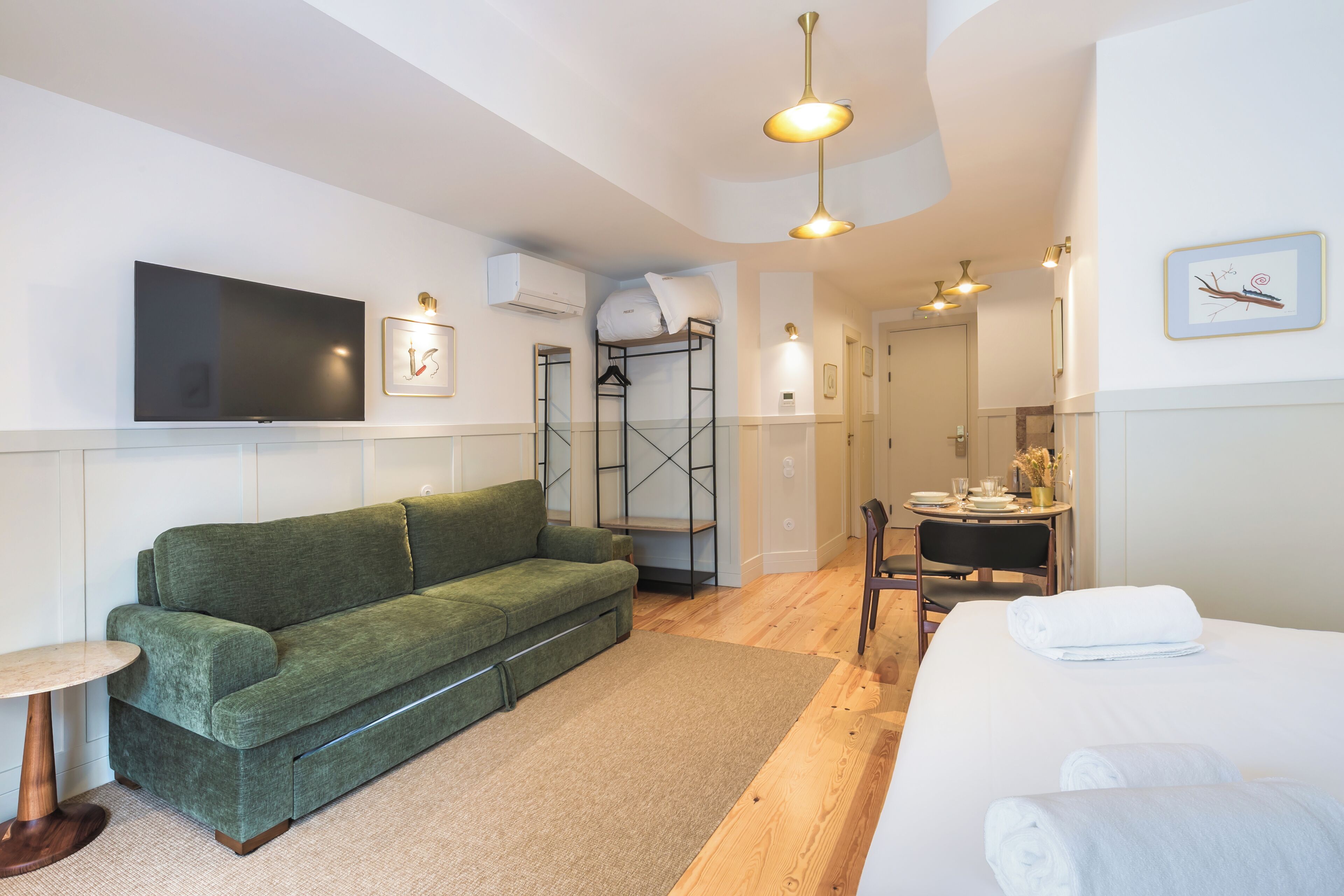 Une chambre d’hôtel moderne avec un canapé vert, un lit avec du linge de lit blanc, une petite table et un téléviseur fixé au mur.