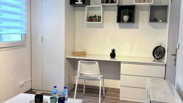 A compact, modern room with a white desk, a chair, a small table with a water bottle, and a cabinet. Shelves with decorative items are mounted on the wall. - Charming apartment in Arras for a cozy getaway (Arras)