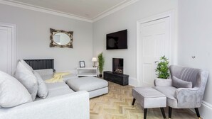 A modern living room with a grey sofa, a grey armchair, a black entertainment unit, a white radiator, and a herringbone-patterned floor. - The Bay Suite - Donnini Apartments (Troon)
