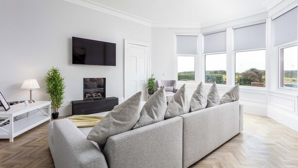 A modern living room with a grey sofa, a flat-screen TV, a white console table, and a large window with a view of greenery. - The Bay Suite - Donnini Apartments (Troon)