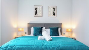 A neatly made bed with a teal duvet, two pillows, and a rolled towel. There are two bedside lamps on either side of the bed. - The Bay Suite - Donnini Apartments (Troon)