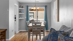 Un comedor con mesa y sillas de madera, una lámpara colgante y vistas al exterior a través de una ventana con cortinas.