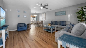 Un amplio salón con un sofá gris, un sillón azul y una mesa de centro. Se ve un ventilador de techo y un letrero que dice "THE BEACH".