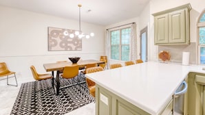 A modern kitchen with a large island, green cabinets, and a dining area with a patterned rug and yellow chairs.