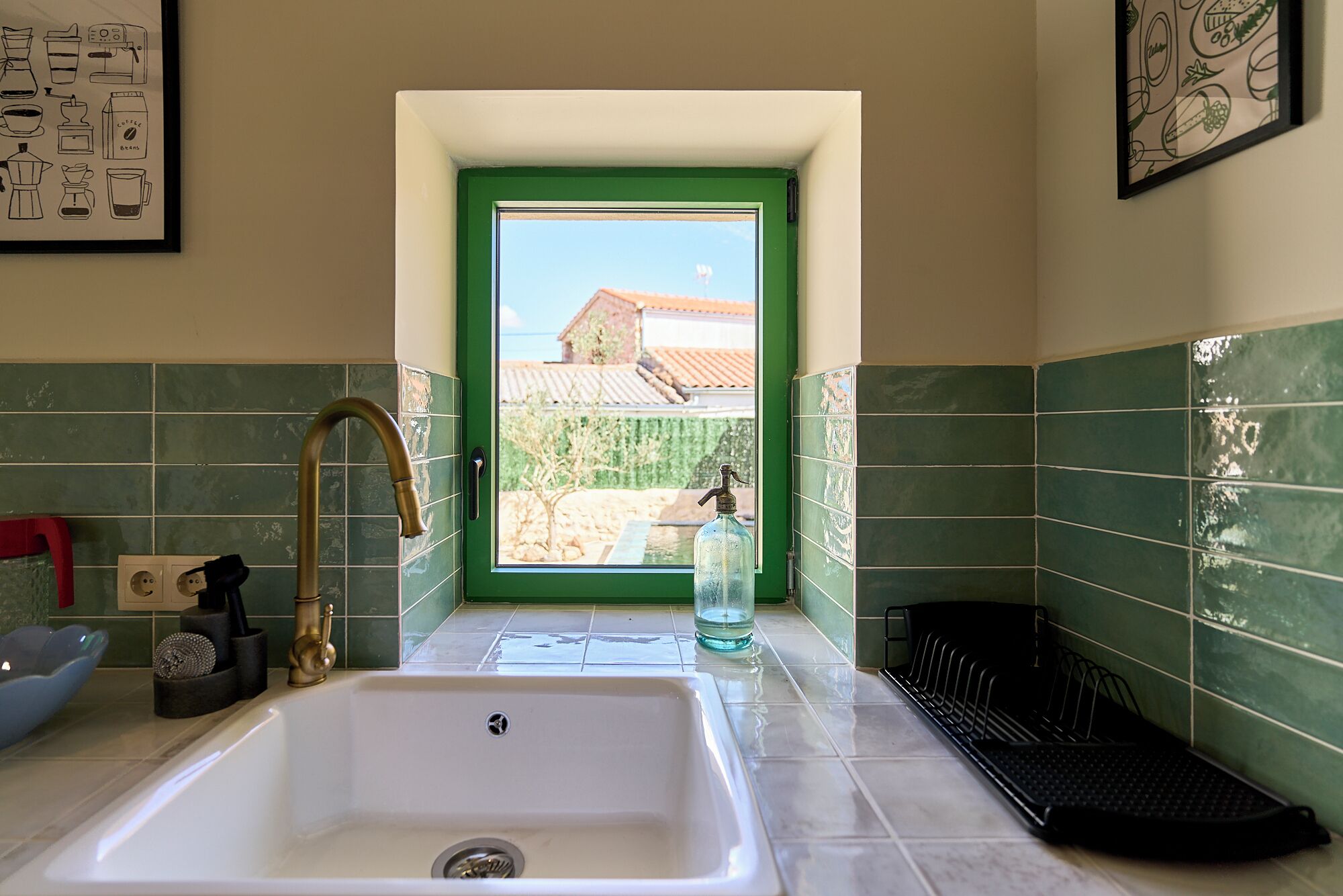 A kitchen with a sink, green tiles, a window, and a view of a house.