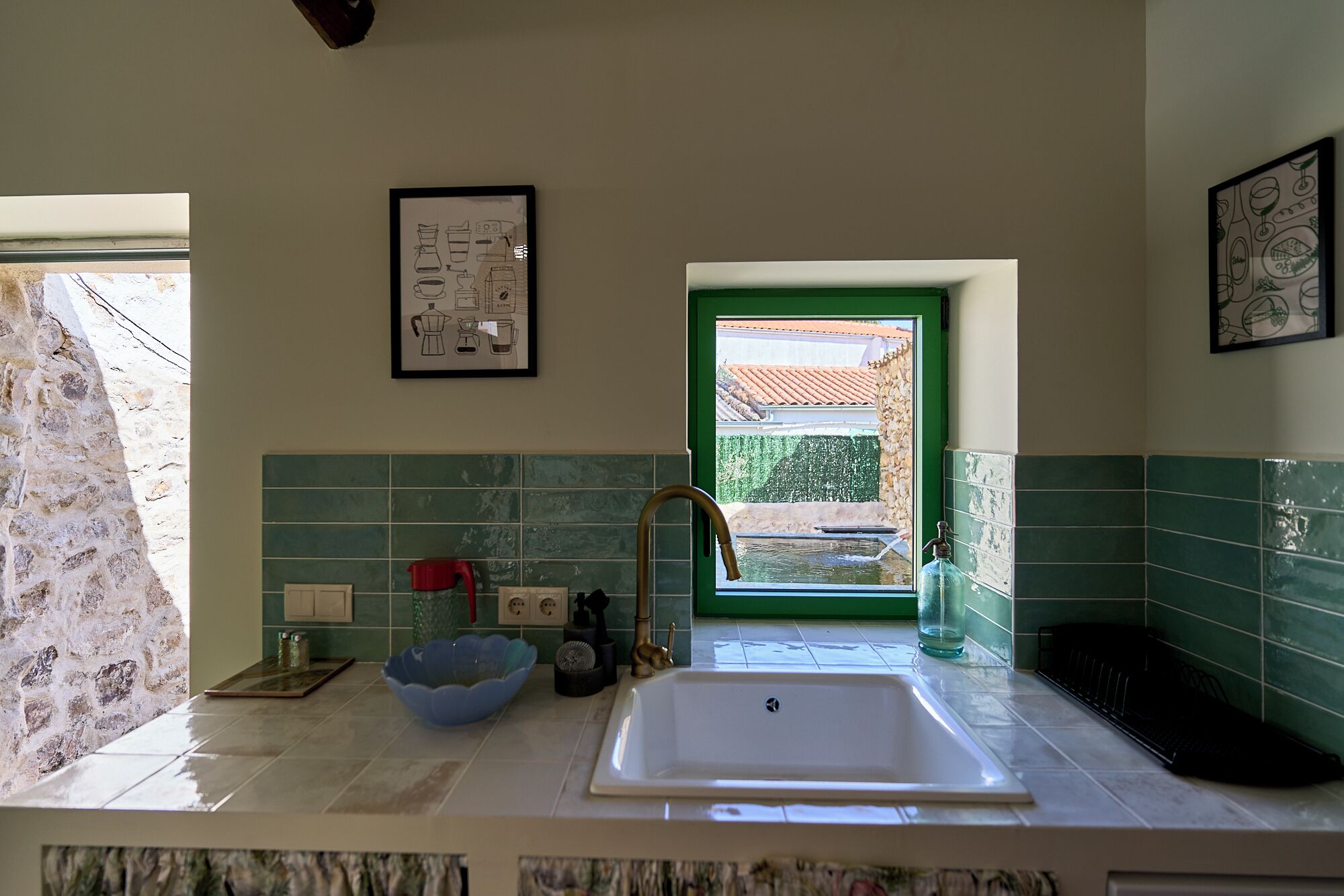 A kitchen with a sink, a window with a green frame, and a tiled backsplash.