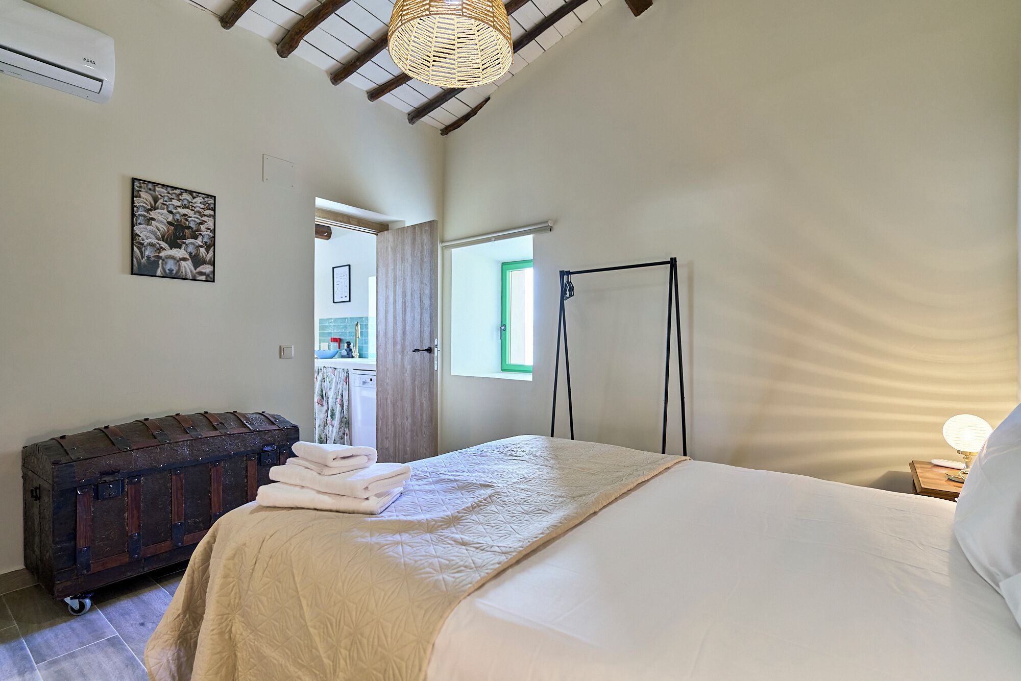 Un dormitorio con cama, maleta, armario, ventana y puerta que da al baño.