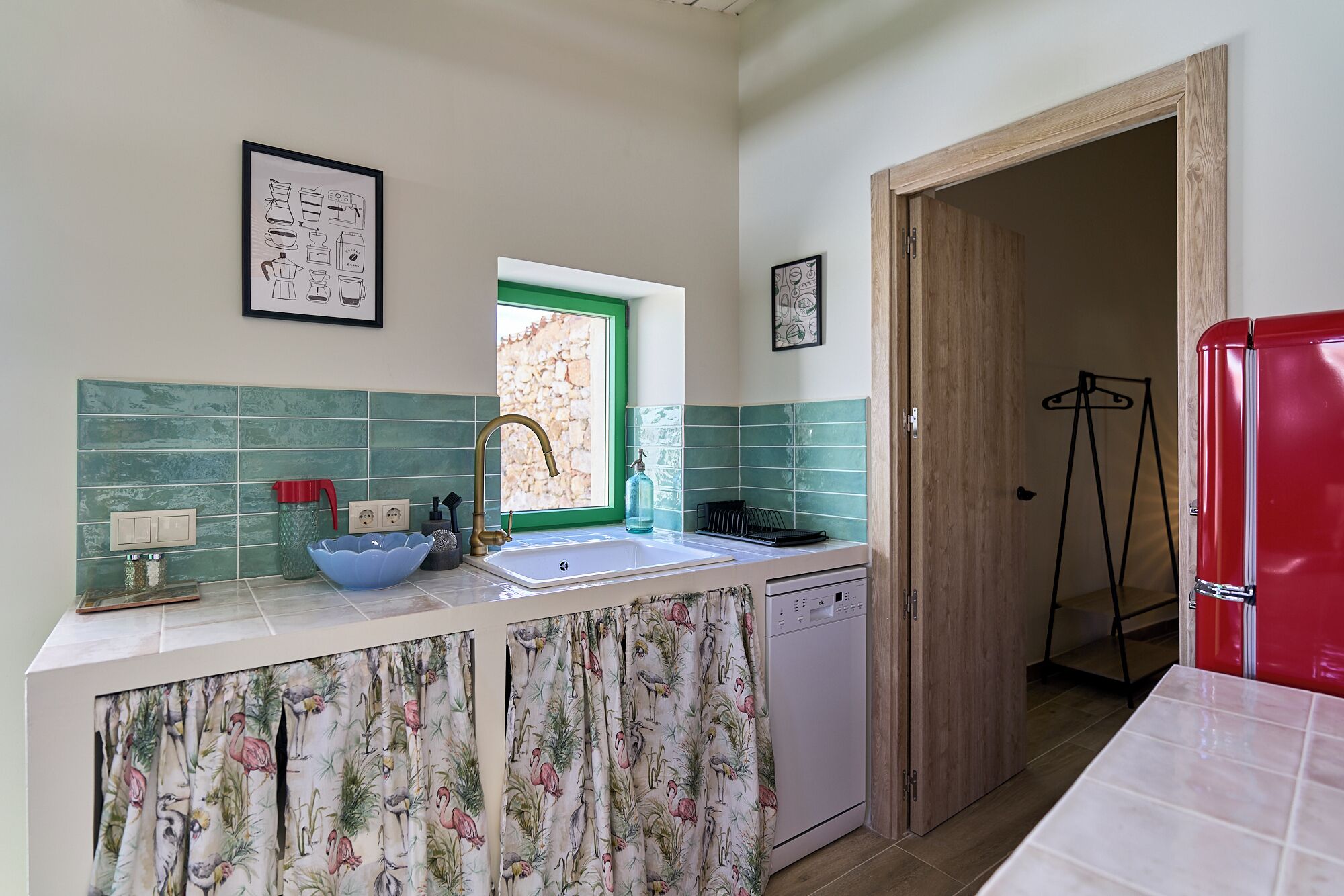A kitchen with a sink, a dish rack, a red refrigerator, and a window with a green frame.