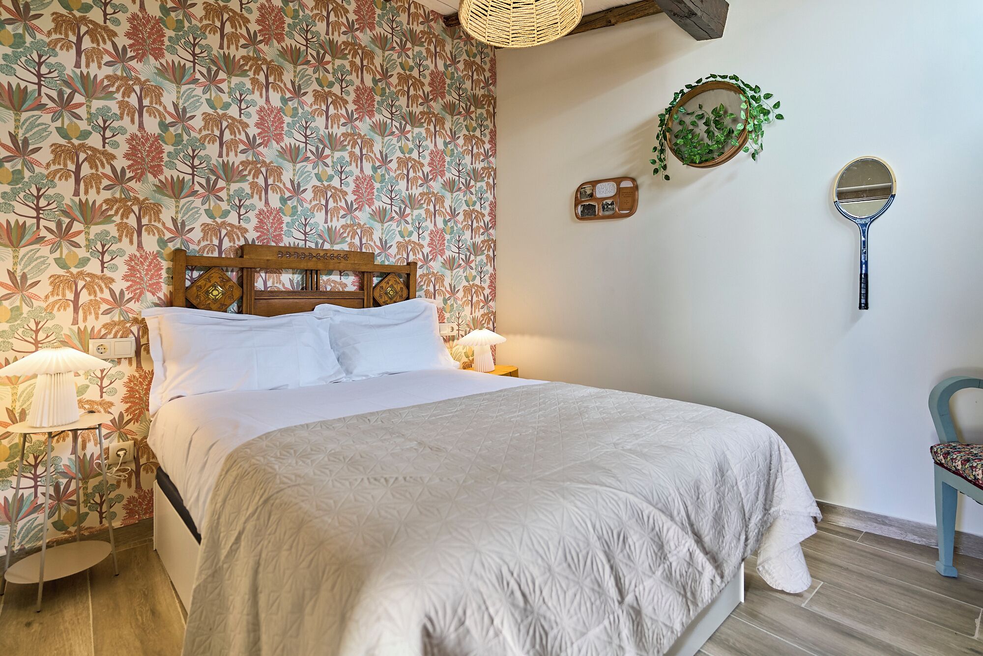 Een net opgemaakt bed met een houten hoofdbord, twee bedlampen, een klein tafeltje met een lamp en een stoel met een bloemenkussen.