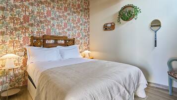 Een net opgemaakt bed met een houten hoofdbord, twee bedlampen, een klein tafeltje met een lamp en een stoel met een bloemenkussen.