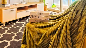 Una sala moderna con un gran ventanal, un televisor de pantalla plana, un mueble bajo de madera, una planta verde y una pila de libros.