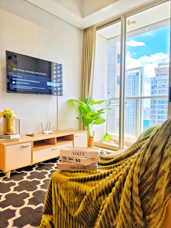 A modern living room with a flat-screen TV, a plant, a bookshelf, and a window with a city view. - Staysanliving - Taman Anggrek Residence (Jakarta)
