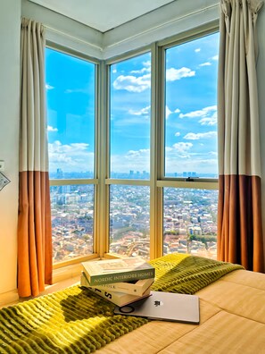 A room with a large window offering a city view, a bed with a green blanket, and a laptop on the bed.