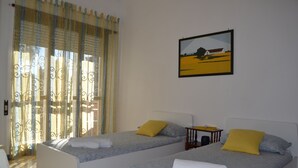 Una habitación con dos camas individuales, cada una con una almohada amarilla y una toalla blanca. Se observa un cuadro en la pared y una ventana con cortinas.