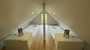 A room with two beds, each with a bedside table and lamp, in a simple, modern design with wooden flooring and a sloped ceiling.