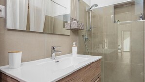 Ein modernes Badezimmer mit einem großen Spiegel, einem weißen Waschbecken, einer Holzkommode und einer Duschkabine aus Glas.