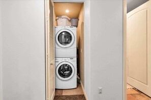 A laundry room with two stacked front-loading washing machines and a small shelf with laundry baskets. - Desert Adobe A | 1BR Legacy Villa (La Quinta)